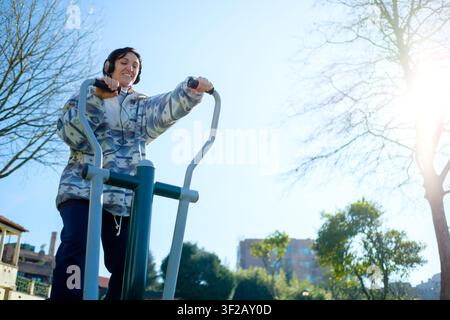 Woman with headphones exercising on an elliptical machine in a public park, enjoying a sunny day Stock Photo