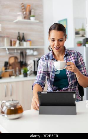 Woman smiling using tablet pc in the morning and enjoying a cup of hot green tea, Working from home using device with internet technology, typing, on gadget during breakfast. Stock Photo