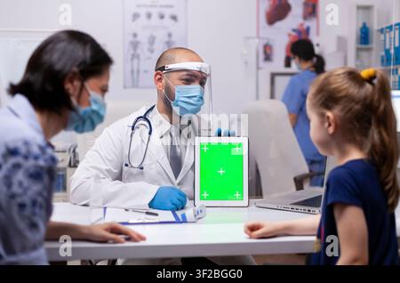 Pediatrician uses a green screen tablet during examination in a medical office while wearing protective gloves and mask, inside a healthcare consultation setting. Stock Photo
