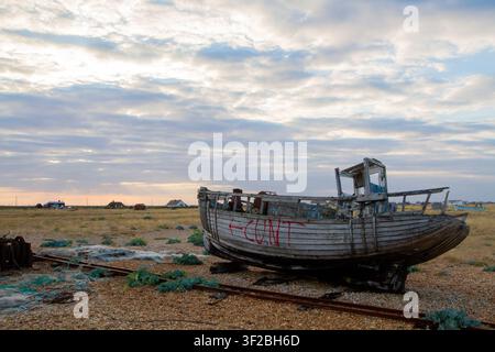 Derelict wooden fishing boat daubed with expletive graffiti beside rusting winch rails and discarded nets on the shingle at Dungeness, Kent, England, Stock Photo
