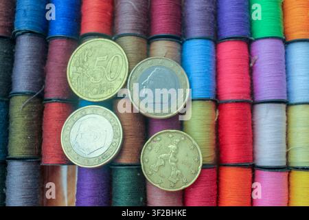 Close up diagonal view of various Euro coins resting on bright colored sewing threads highlighting the increasing prices of basic goods, craft materia Stock Photo