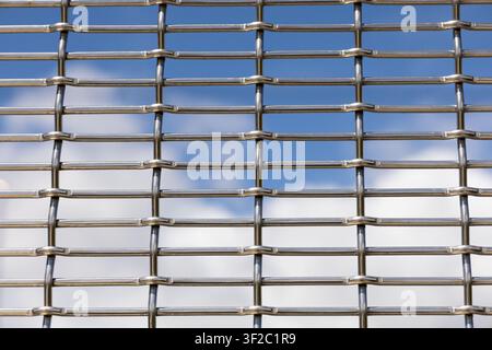 Close-up of woven stainless steel wire mesh with a blue sky and soft clouds visible through the openings. Stock Photo