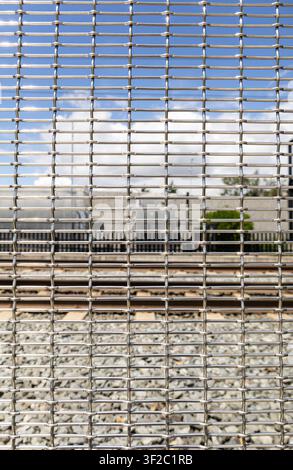 Close-up of woven steel wire mesh fence with railway tracks and an industrial background visible through the openings. Stock Photo