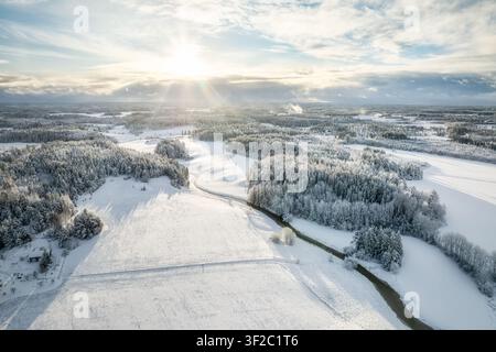 Aerial view of a snow-covered winter landscape with dense pine forests and a winding river under a bright sunburst in Lieto, Southwest Finland, Finlan Stock Photo
