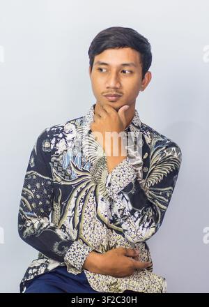 Thoughtful Asian man wearing traditional batik shirt thinking deeply with hand on chin isolated on studio background Stock Photo