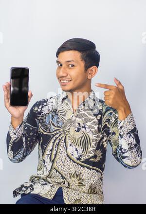Asian man wearing traditional batik shirt pointing at blank smartphone screen mockup isolated on studio background Stock Photo