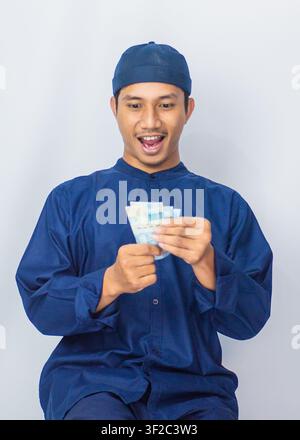 Happy Indonesian man holding banknote money cash with joy celebrating financial success in studio. Stock Photo
