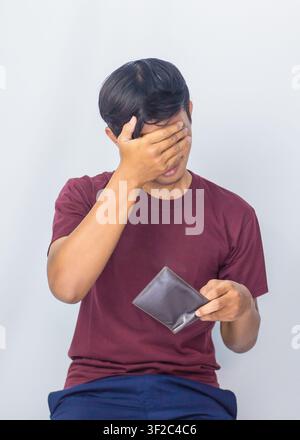Disappointed Indonesian man covering his face with empty wallet feeling broke and sad in studio. Stock Photo