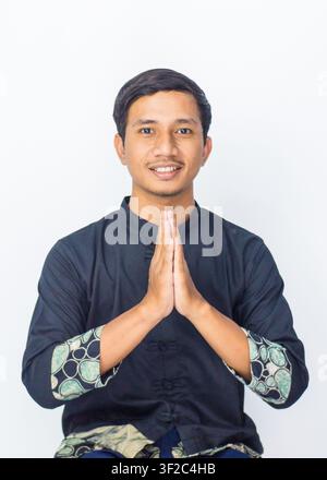 Smiling Asian male model wearing traditional shirt showing respectful greeting posture on blue studio background. Stock Photo
