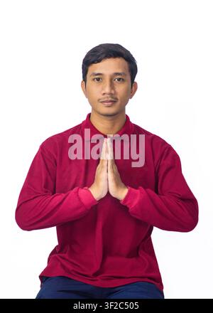Smiling young Asian man in red traditional shirt offering polite Indonesian greeting gesture isolated on white. Stock Photo