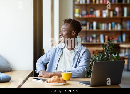 Smiling black guy using laptop computer at cozy cafe, having hot drink with croissant, looking out window Stock Photo