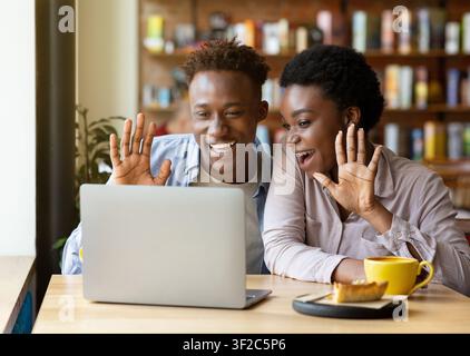 Young black couple communicating online on laptop computer at cozy urban cafe, waving to camera, greeting someone Stock Photo