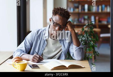 Focused African American student taking notes from book in cafe Stock Photo