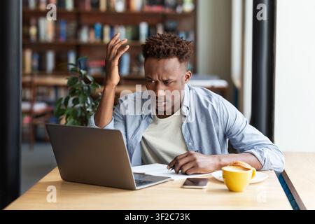 Irritated African American guy using laptop to work on difficult business project at city cafe Stock Photo