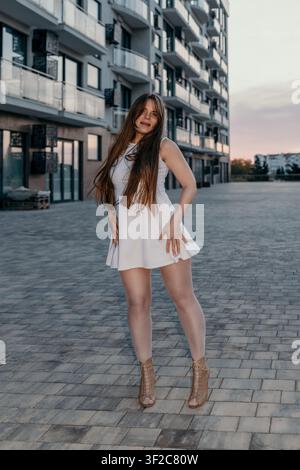 Woman fashion city posing in white dress near modern residential buildings during evening sunset light Stock Photo