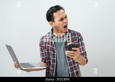 Angry mad asian indonesian man holding smartphone and laptop computer on isolated background Stock Photo