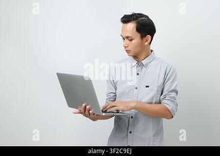 Pensive young Asian Indonesian man holding laptop computer with a serious face thinking about a question, thoughtful about confusing idea on isolated Stock Photo