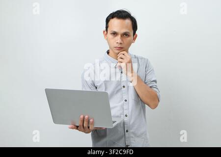 Pensive young Asian Indonesian man holding laptop computer with a serious face thinking about a question, thoughtful about confusing idea on isolated Stock Photo