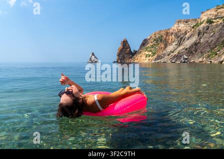 Woman float sea floating on a pink inflatable ring in crystal clear water near rocky cliffs during summer vacation for relaxation Stock Photo