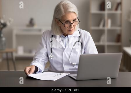 Focused senior woman doctor do paperwork at workplace using laptop Stock Photo