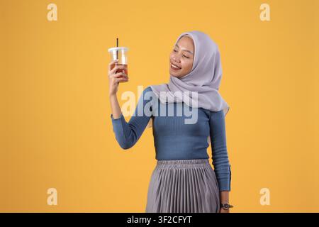 happy asian indonesian muslim woman holding and drinking ice tea in plastic cup on isolated yellow background Stock Photo