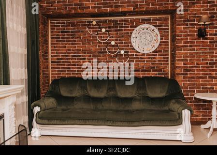 Elegant living room interior with a green velvet sofa, decorative brick wall, vintage clock, and warm ambient lighting. Cozy classic home design with stylish furniture and luxurious decor, perfect for interior design and real estate concepts. Stock Photo