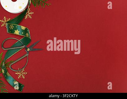 Paper spool of green Christmas ribbon with golden snowflakes, scissors and green evergreen twigs for holiday gift wrapping on red paper background Stock Photo