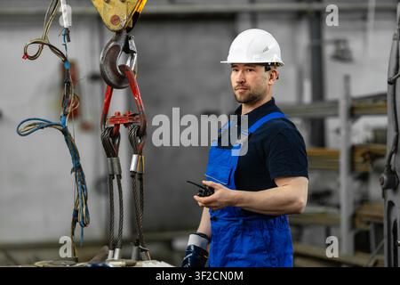Industrial engineer with two-way radio standing near overhead crane hook in factory Stock Photo