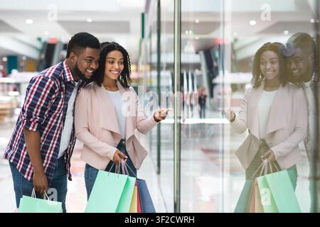 Happy Black Spouses Enjoying Shopping Together In Mall, Looking At Showcase Stock Photo