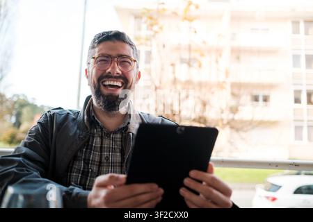 Man with beard and eyeglasses laughing while using a digital tablet on a city balcony Stock Photo