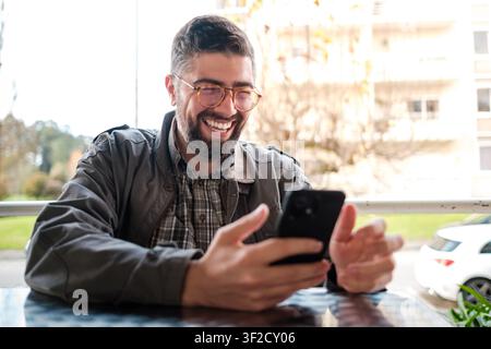 Cheerful man with glasses enjoys smartphone browsing at a sunny outdoor café Stock Photo
