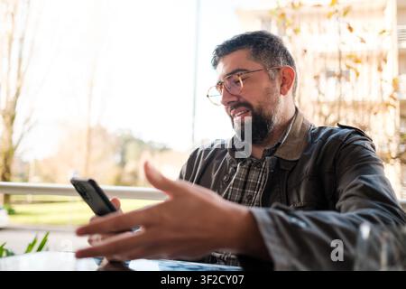 Man actively talking on his smartphone, gesturing with hands, sitting at an outdoor table Stock Photo