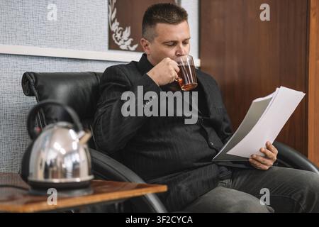 Businessman in a black suit sitting in a leather armchair, drinking hot tea from a glass mug while reading paperwork. Office break, daily work routine Stock Photo