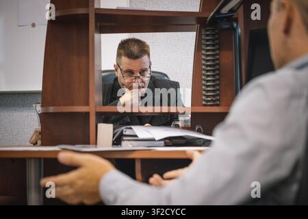 Thoughtful boss wearing glasses and touching his chin, listening to a colleague across the desk. Stock Photo