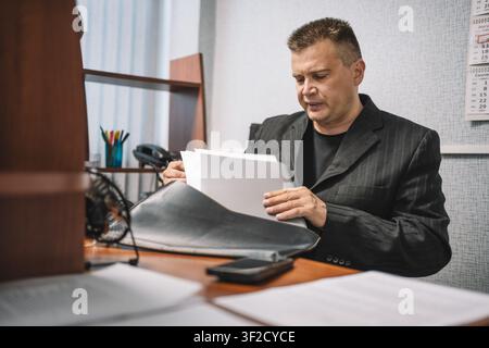 Mature businessman in a dark suit reading and analyzing documents while sitting at office desk in a professional corporate environment. Stock Photo