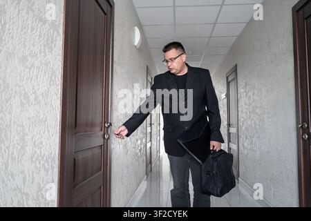 Man with glasses and a briefcase using a key to open an office door in modern office corridor. Stock Photo