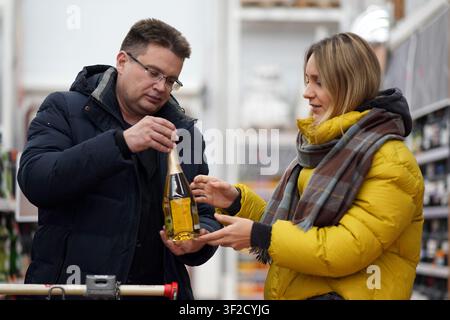 Smiling man holding a bottle of sparkling wine while talking to a woman in a yellow jacket during grocery shopping trip. a man chooses wine in a alcoh Stock Photo