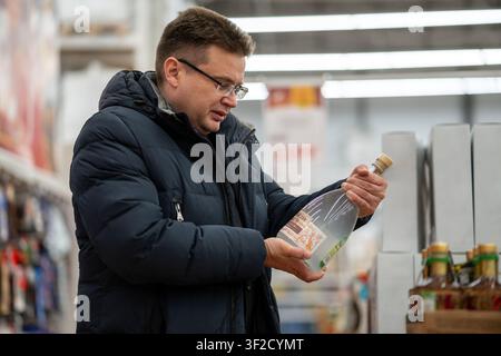 Man in a blue jacket reading the label on a large transparent bottle while standing in the grocery aisle of a store. Stock Photo