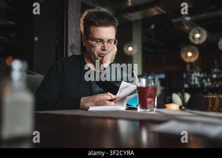 Man with glasses reading an open book while resting his chin on his hand, with a glass of red drink on the table. Stock Photo
