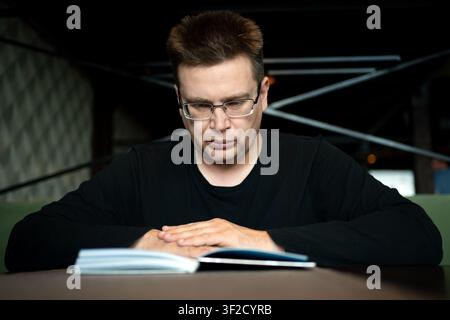 Man with glasses sitting at a dark table, looking down at an open book with his hands resting peacefully on it. Stock Photo