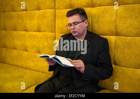 Man with glasses sitting on a bright yellow tufted sofa reading an open book in a modern interior. Stock Photo