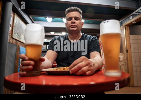 Tired man in a dark T-shirt drinking beer from large glass mug with white foam, sitting at a bar table. Stock Photo