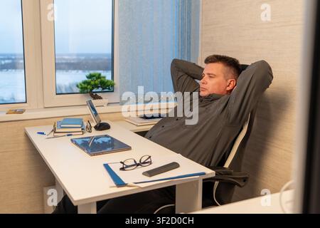 Man relaxing in an office chair with hands behind his head, sitting at a white desk next to window with a bonsai tree. fatigue at the end of the worki Stock Photo