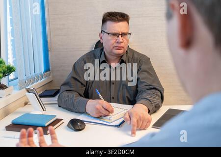 Businessman interviewing a job candidate while taking notes during an office meeting, suitable for recruitment, journalism, business interview, or pol Stock Photo
