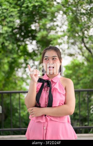 A vertical medium shot of a young Southeast Asian Thai Isan woman in a pink top with a black bow ribbon, looking away with an open mouth and pointing Stock Photo