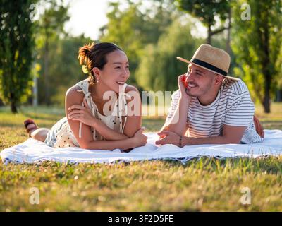 Two friends lie on a blanket in a sunlit park, sharing laughter and conversation during a relaxing picnic. Stock Photo