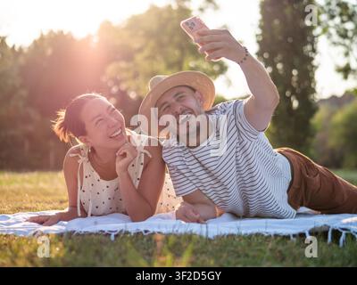Multiracial couple spending time together in park in summertime. They are using smartphone and taking selfie. Stock Photo