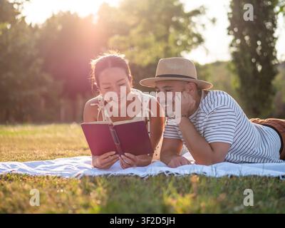 Happy multiracial couple is spending time together in park in summertime. They are reading book. Stock Photo