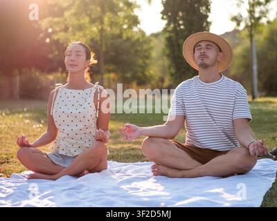 Adult multiracial couple is practicing yoga in park in summertime. Lotus position. Stock Photo