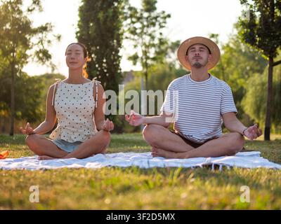 Adult multiracial couple is practicing yoga in park in summertime. Lotus position. Stock Photo